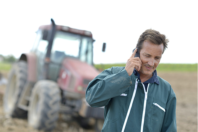 A farmer chatting on his phone with a tractor behind him on a field.