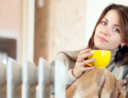 A lady sits in a blanket with a yellow cup near a radiator