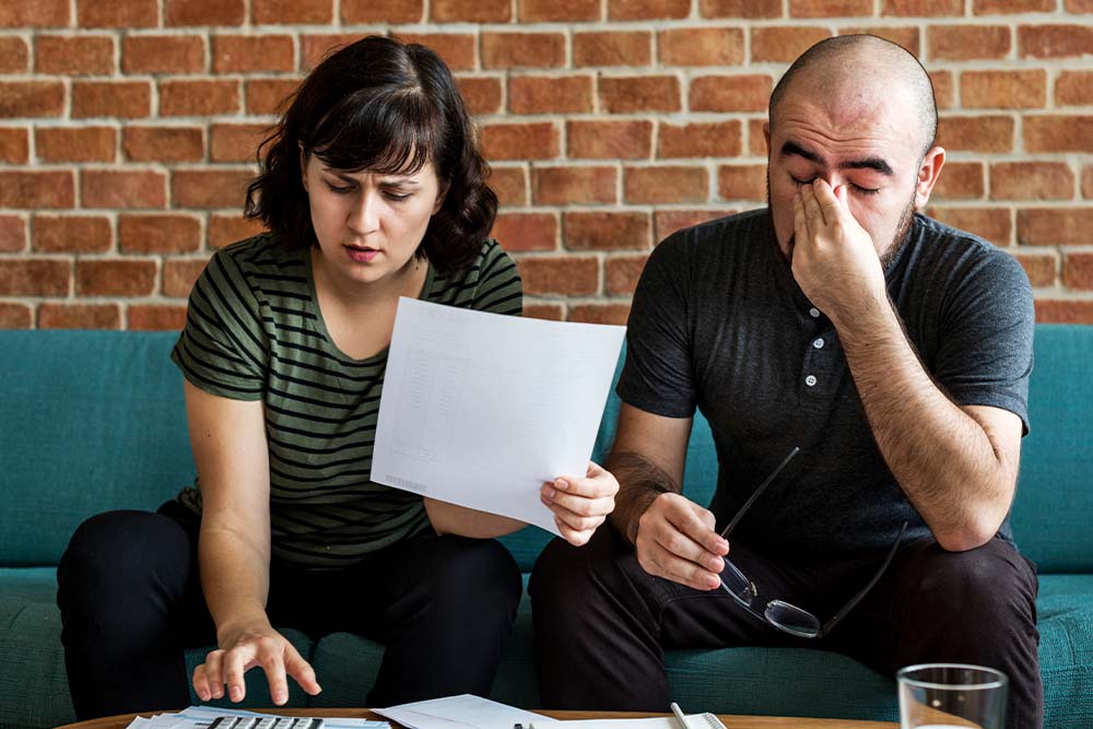 A man pinches his eyes as a woman next to him holds a paper as they work out debt