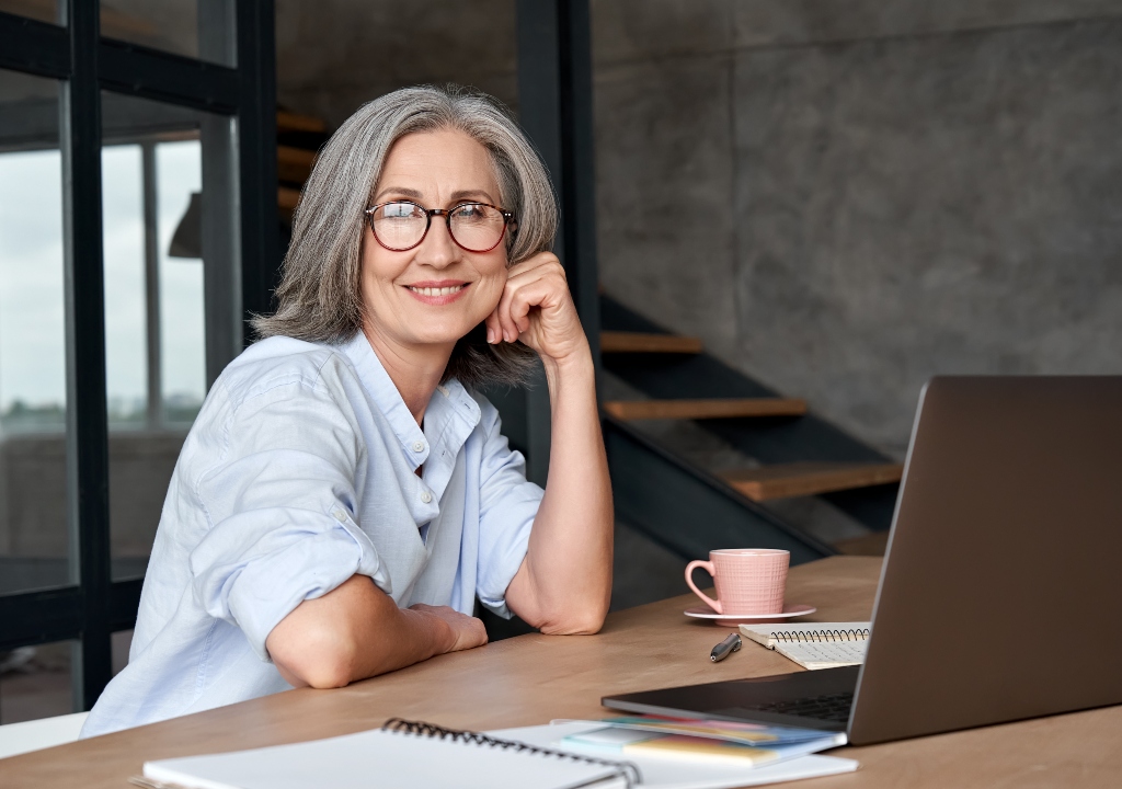 Smiling stylish mature middle aged woman sits at desk with laptop