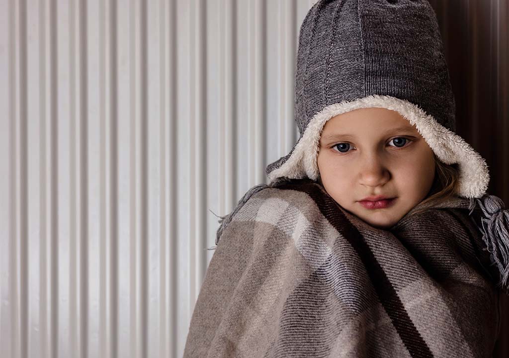 A child dressed in warm clothes inside leaning against a radiator to stay warm.