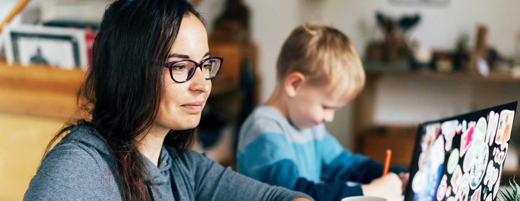 A woman sits with her laptop next to a child who is drawing.